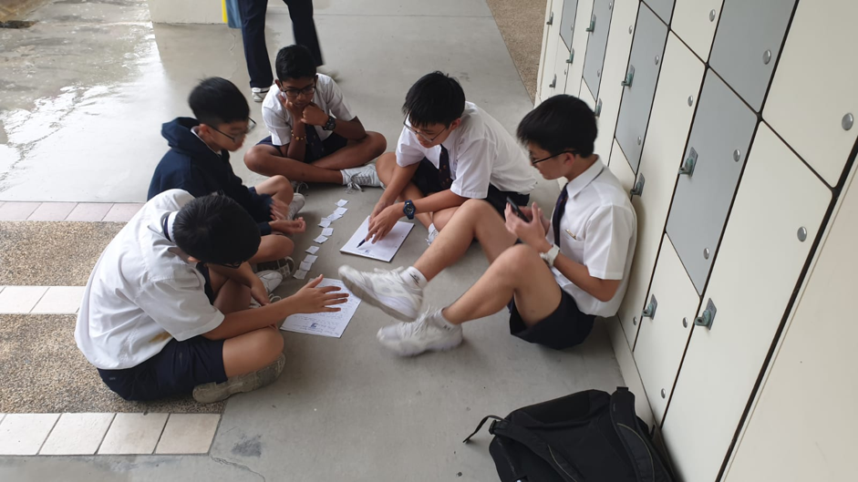 A small group of students in school uniforms sit together on the floor in a corridor, gathered around papers or worksheets, appearing to collaborate or study together.